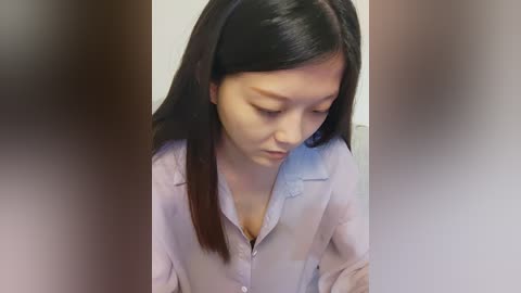 Media: A video of an Asian woman with long black hair, wearing a light purple button-up shirt, leaning forward, looking down, with blurred background.