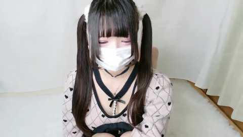 Media: Video of an Asian woman with long, dark hair in twin pigtails, wearing a white face mask, a black choker, and a white dress with black polka dots, kneeling on a white floor.