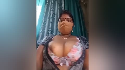 Media: Video of a South Asian woman with medium skin tone, wearing a floral-patterned bra, a face mask, and glasses, standing in a room with teal curtains.