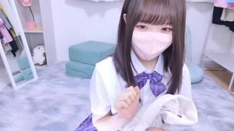 Media: A video of an Asian girl with straight, dark brown hair and pale skin, wearing a white blouse, purple tie, and face mask, sitting on a light gray carpet in a brightly lit room with teal furniture.