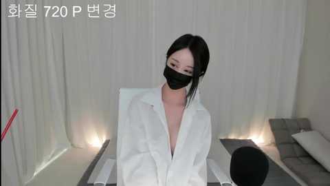 Media: A video of an East Asian woman with fair skin and long black hair, wearing a white blouse, black mask, and glasses, seated in a minimalist room with white curtains and a grey couch.