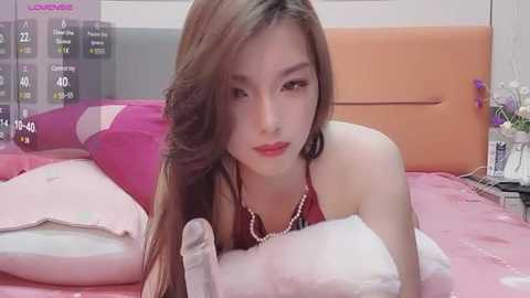 Media: Video of an Asian woman with long, straight brown hair, fair skin, wearing a red halter top and pearl necklace, lying on a pink bed, surrounded by plush pillows.