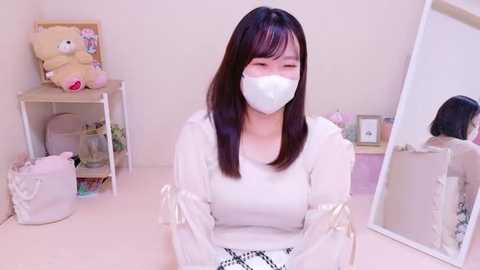 Media: Video of an East Asian woman with straight black hair, wearing a white face mask and long-sleeved white blouse, sitting at a white desk in a light-colored room filled with teddy bears, a mirror, and decorative items.