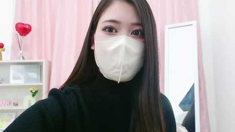 Media: Video of a young Asian woman with long black hair, wearing a white surgical mask, black turtleneck sweater, and a pink room with a heart-shaped red vase and a white shelf.