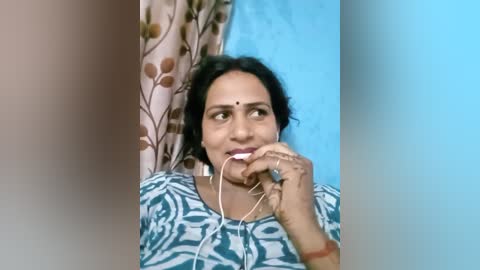 Media: Video of an Indian woman with medium brown skin, dark hair, and a small bindi, wearing a blue and white patterned blouse. She holds white earbuds in her mouth, smiling, against a backdrop of floral curtains and a blue wall.