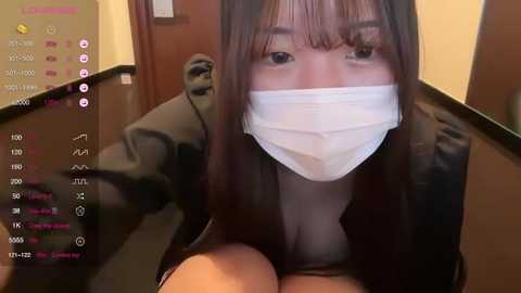 Media: A video of an Asian woman with long black hair, wearing a white face mask, seated on a black chair in a dimly lit room.