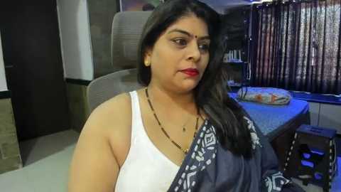 Media: Video of a middle-aged South Asian woman with medium skin tone, wearing a white sleeveless top and dark sari, seated in a modern living room with wooden furniture and vertical blinds.