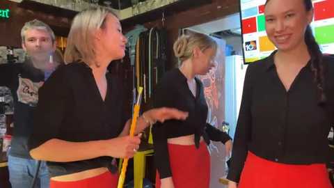 Media: Video of four people in a cluttered, rustic room. Two women in black tops and red skirts, one with a yellow paddle, interact with a bearded man and a woman with long hair. Background includes shelves and hanging items.