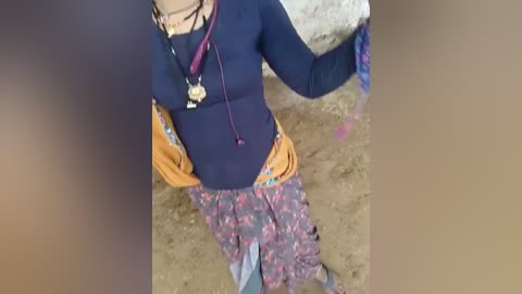 Media: A video showing a woman from the waist up, wearing a blue long-sleeve shirt, multi-colored patterned skirt, and carrying a tan bag. Background features dirt ground and a blurred person.