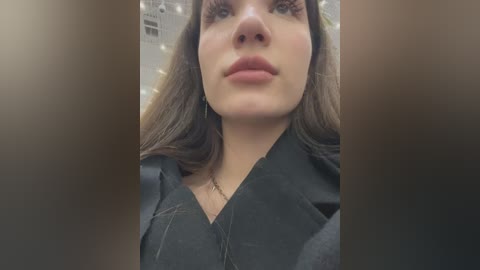 Media: Video of a young Caucasian woman with fair skin, long brown hair, and a neutral expression, wearing a black shirt. The blurred background suggests an indoor setting with soft lighting.