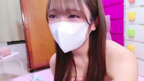 Media: Video of an Asian woman with long brown hair, wearing a white surgical mask, in a brightly colored room with a pink wall and yellow pegboard.