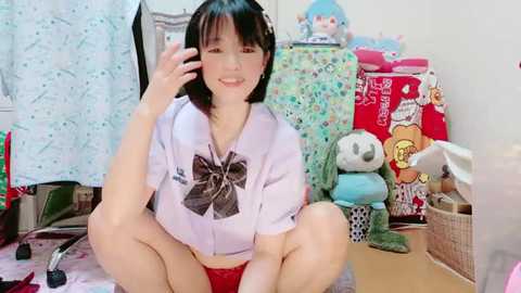 Media: Video of an Asian woman with short black hair, wearing a light purple shirt with a black bow, squatting on a pink carpet. Background features stuffed toys and a red suitcase.