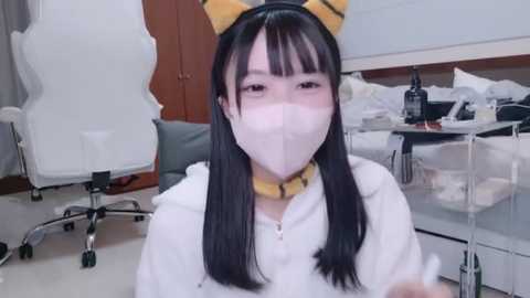 Media: Video of an Asian woman with long black hair and yellow cat ears, wearing a white face mask and a white hoodie, sitting in a cluttered room with medical equipment and a bed.