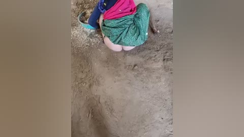 Media: A video captures a person in a green and pink skirt, kneeling on a muddy ground, holding a blue bucket. The person's legs and buttocks are visible.