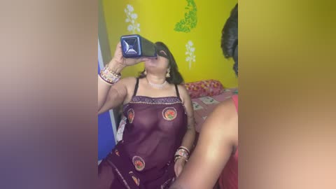 Media: Video of a dark-skinned woman in a sheer maroon nightgown with floral patterns, sitting on a bed in a colorful room with yellow walls and green patterns. She is taking a selfie with a smartphone.