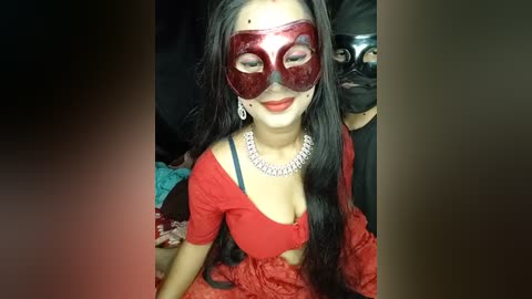 Media: Video of a woman with long black hair, wearing a red Venetian mask, red off-shoulder top, and silver choker, seated on a black couch with a black mask-wearing person partially visible in the background.