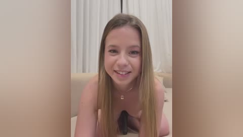 Media: A video of a young, nude, fair-skinned woman with long, straight, blonde hair, kneeling on a beige bed. She has a cheerful expression, wearing a delicate necklace. The background features white curtains and a beige pillow.