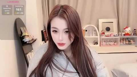 Media: Video of an Asian woman with long brown hair, fair skin, and light makeup, wearing a light grey jacket, sitting in a bedroom with a mirror, shelves, and stuffed animals.