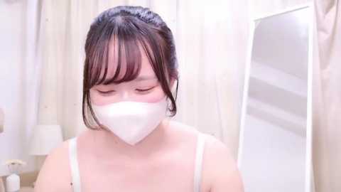 Media: Video of a young Asian woman with fair skin, wearing a white face mask, white spaghetti-strap top, and black hair tied back. She stands indoors, in front of a mirror and white curtains, in a bright, softly lit room.