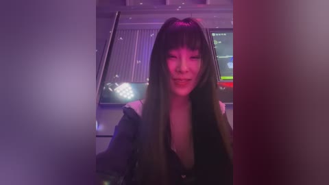 Media: Video of a smiling, light-skinned woman with long, straight black hair, wearing a black top, against a purple-lit background with a computer screen visible.