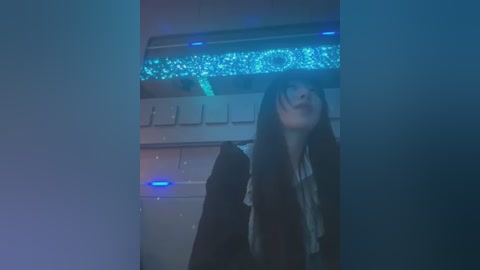 Media: Video of a young woman with long, dark hair and pale skin, wearing a dark coat, standing in a dimly lit, futuristic room with glowing blue LED lights and metallic textures.