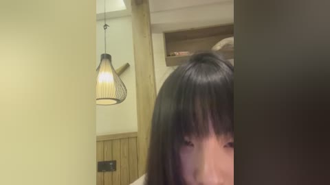 Media: Video of a woman with long black hair partially covering her face, standing in a modern, minimalist room with wooden paneling, a hanging lamp, and a shelf in the background.