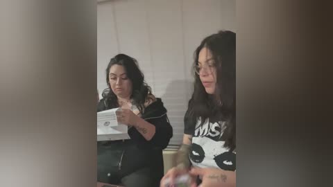 Media: Video of two women, one with long dark hair, wearing a black hoodie, sitting in a dimly lit room, reading a document. Another woman with long dark hair, wearing a panda-themed t-shirt, is standing in the foreground.