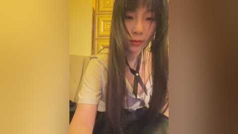 Media: Video of an Asian woman with long, straight black hair, wearing a white blouse and black suspenders, standing in a dimly lit room with golden geometric patterns on the wall.