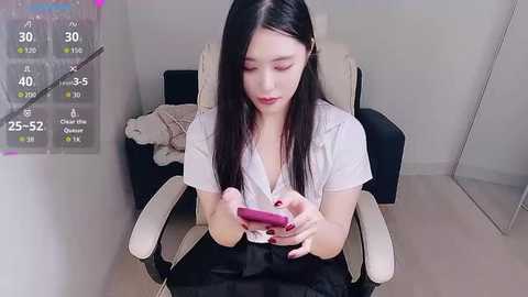 Video of an Asian woman with long black hair, seated in a chair, holding a pink vibrator, in a minimalist room.