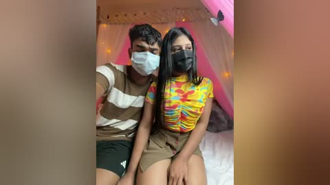 Video of a young couple sitting close, wearing face masks, the woman in a colorful top, the man in a striped shirt. Background includes a bed with a pink canopy and string lights.