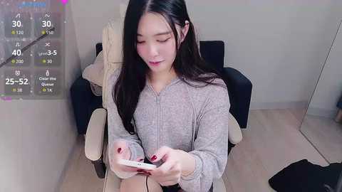 Media: A video of an Asian woman with long black hair, light skin, and red-painted nails, sitting in a beige armchair, playing a smartphone game.