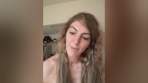 Video of a slender, fair-skinned woman with long, wavy, light brown hair, wearing no clothing, standing in a dimly lit, cluttered room with a closet visible in the background.