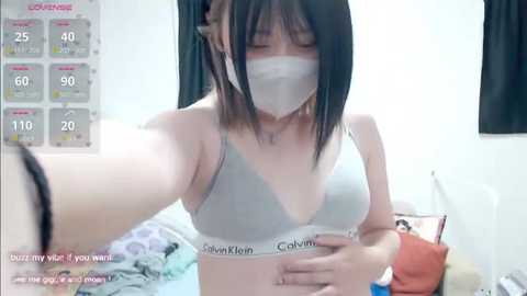 Video of an Asian woman with short black hair, wearing a gray Calvin Klein bra and face mask, taking a selfie in a cluttered bedroom.