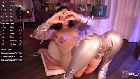 Media: Video of a woman in a silver, skin-tight bodysuit, lying on a pink cushion, making a heart shape with her hands. Background shows a dimly lit room with furniture and a digital camera.