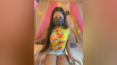 Video of a young woman with long dark hair, wearing a face mask, colorful tie-dye top, and denim shorts, seated on a bed with pink and yellow draped canopy, in a cozy, softly lit bedroom.