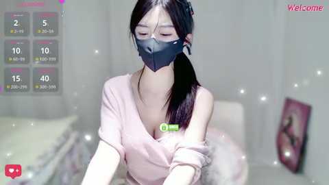 A young Asian woman with long black hair wears a face mask and pink top in a softly lit bedroom, with a digital clock and \"Welcome\" text in the top right corner.