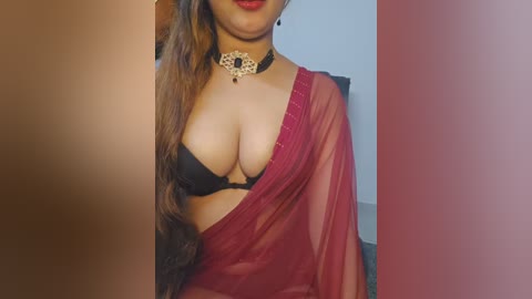 Video of a South Asian woman with long brown hair, wearing a black bra, red sari, and a gold choker. The background is blurred, with a beige wall.