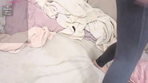 Media: Video of a disheveled bedroom with a person lying on a bed, partially covered by a blanket, and a pair of legs wearing black pants visible in the foreground.