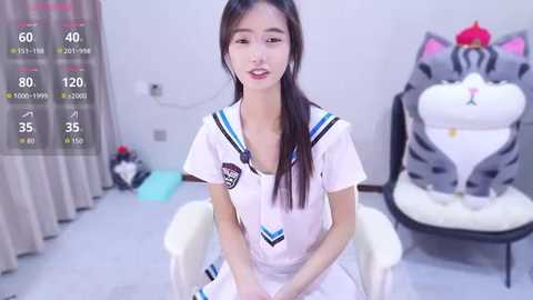 Video of an East Asian woman with long black hair, wearing a white sailor uniform, sitting in a white chair in a minimalist room with a large white teapot and red flowers.
