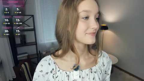 Media: Video of a young Caucasian girl with light brown hair, wearing a white floral top, smiling. Background features a lamp, shelves, and a window.