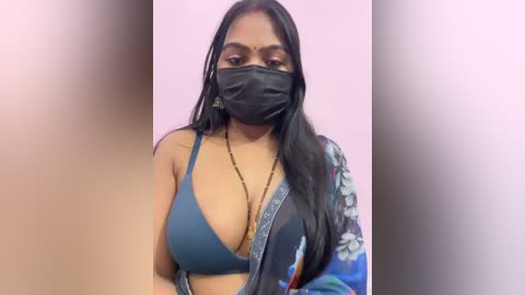 Video of a South Asian woman with long black hair, wearing a blue bra, black face mask, and a floral-patterned shawl. She has medium skin tone and a confident expression. Background is blurred, featuring light pink hues.