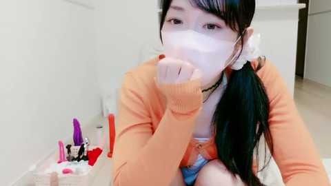 A video of an Asian woman with long black hair, wearing an orange cardigan, mask, and blue underwear, sitting on a white bed, blowing into a pink dildo, surrounded by various sex toys.