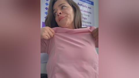 Media: Video of a middle-aged woman with fair skin and brown hair, wearing a pink t-shirt, smiling as she lifts it, in a blurred, pink-tinted office background.