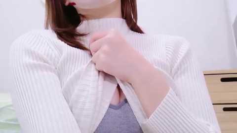 Video of a fair-skinned woman with long, straight brown hair, wearing a white ribbed sweater with a low-cut neckline, exposing a white bra underneath. She is in a white room with a light wooden dresser on the right.
