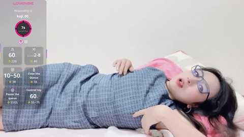 Video of an East Asian woman lying on a bed, wearing a grey checkered dress and glasses, with a virtual health tracking overlay on the left side.