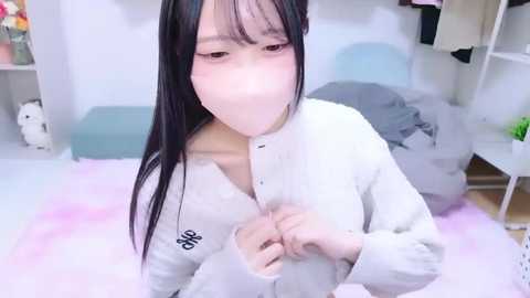 Video of a young Asian woman with long black hair and pale skin, wearing a white button-down shirt, kneeling on a pink rug in a softly lit, minimalistic bedroom.