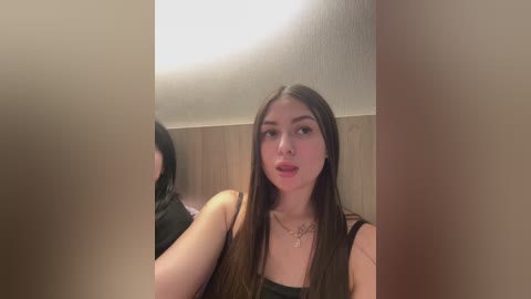 Video of a young woman with long brown hair and fair skin, wearing a black tank top, standing in a dimly lit bathroom with a wooden ceiling.