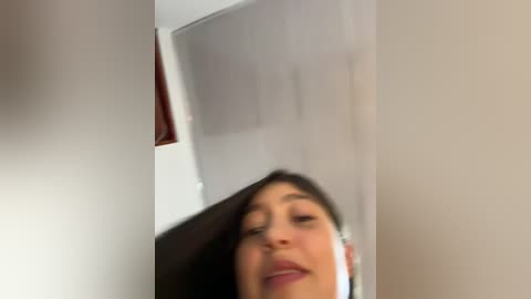 Video of a young woman with straight, dark hair, wearing white earbuds, looking down at a phone, taken from a low angle. Background features white walls and a wooden door.