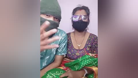 Media: Video of two South Asian individuals, man in green hat and mask, woman in purple floral blouse and green saree, with a blurred background.