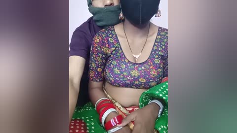 Media: Video of a woman with a dark complexion wearing a colorful floral blouse, a black mask, and traditional jewelry, seated with a man's arm around her.